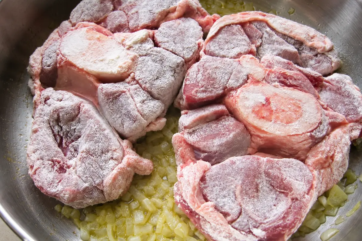 Flour-coated veal shanks in a pan with softened chopped onion.