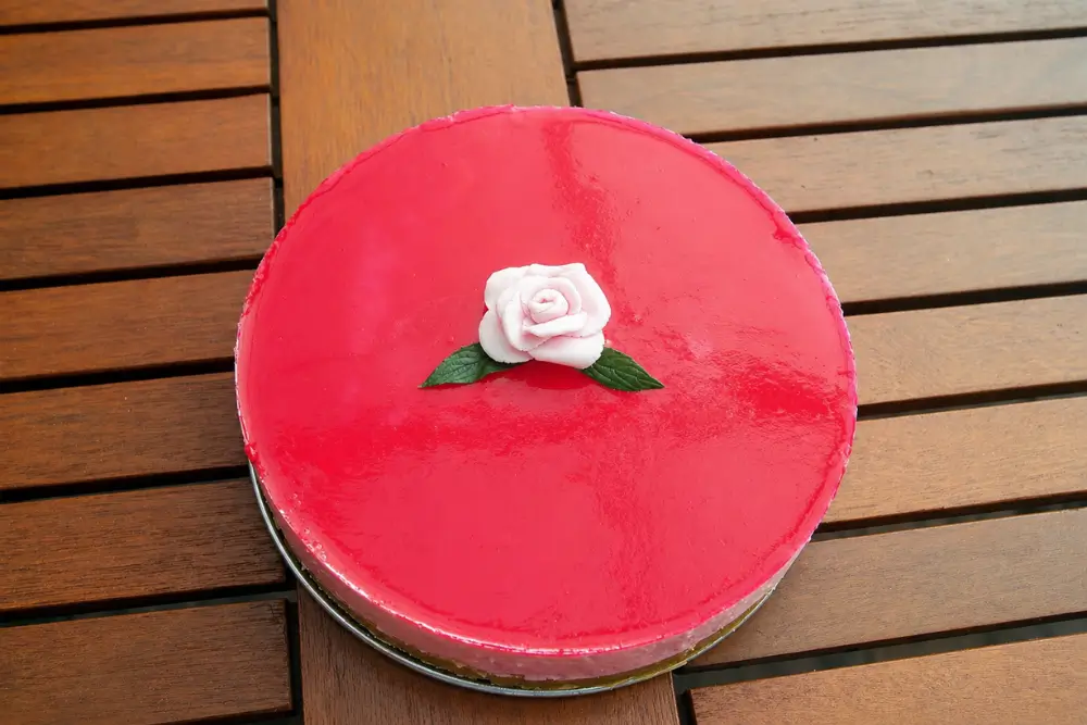 Chilled cheesecake with glossy pink topping and fondant rose decoration on a wooden table.