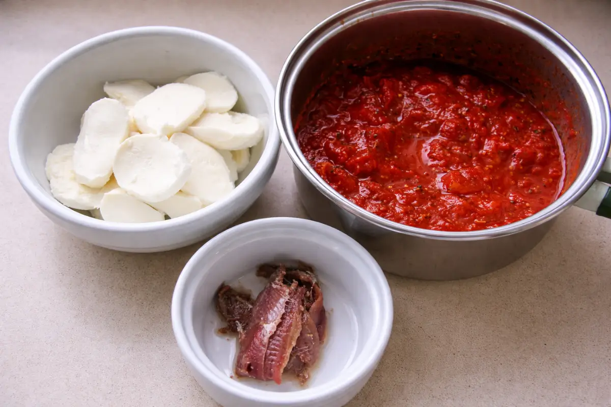 Sliced mozzarella, tomato sauce, and anchovies prepared for topping.