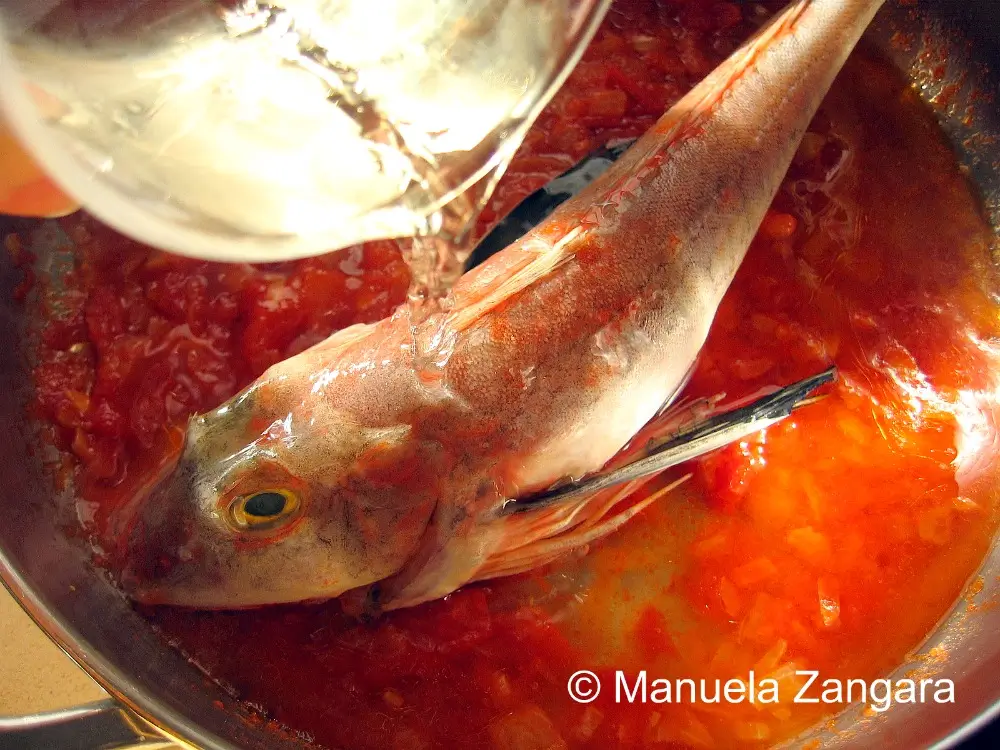 White wine being poured over a whole fish in tomato sauce.