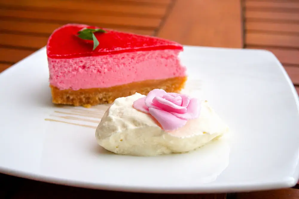 Slice of Rose Cheesecake served with whipped cream and a fondant rose on a white plate.