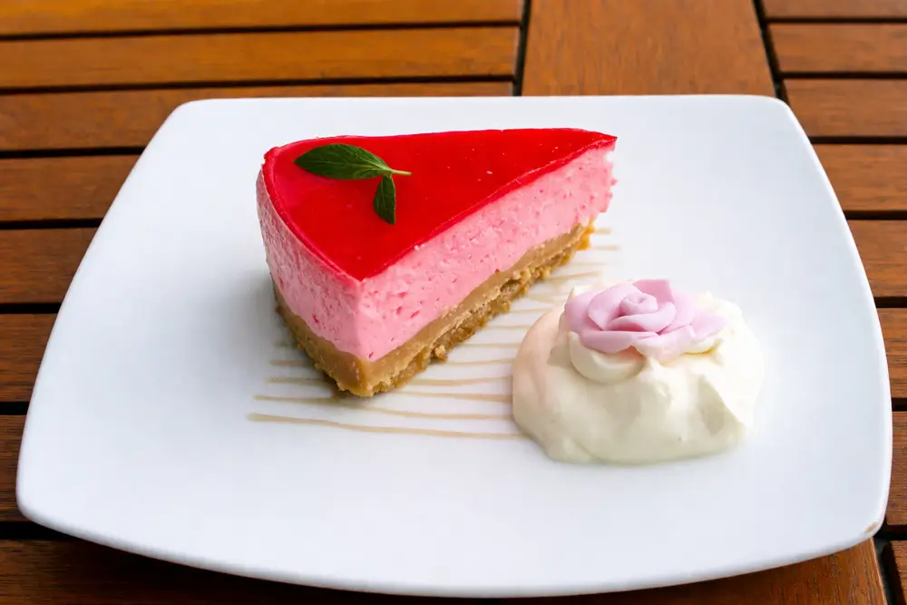 Slice of chilled cheesecake with glossy rose topping served with whipped cream and a fondant rose.