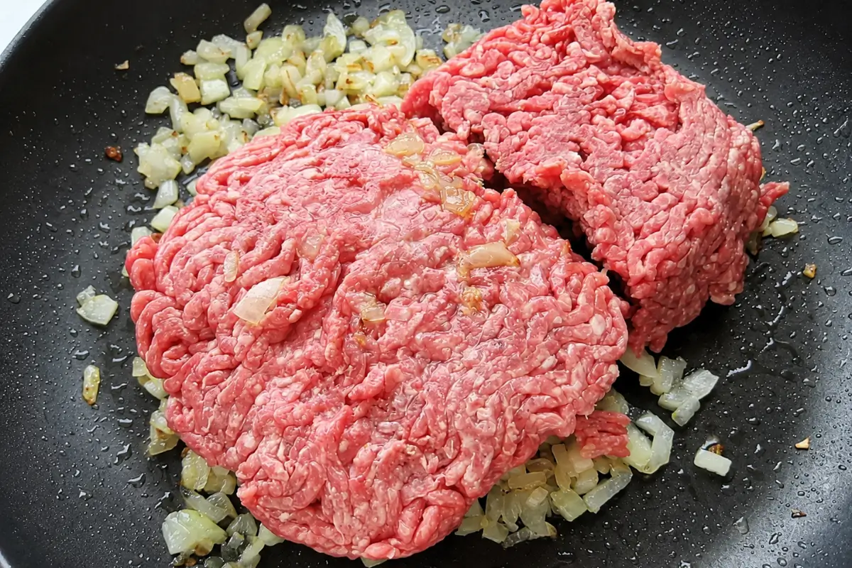 Raw ground beef added to softened onions in a black frying pan.
