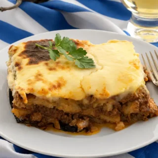 A thick slice of Greek moussaka served with parsley beside a fork and white wine.