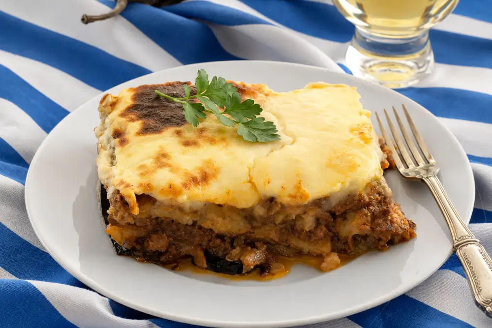 A thick slice of Greek moussaka served with parsley beside a fork and white wine.