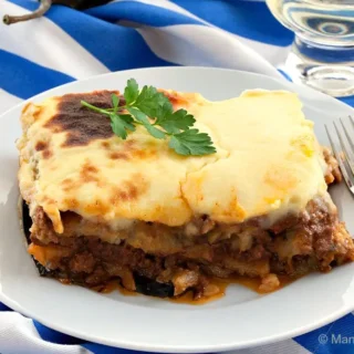 A plated slice of baked moussaka with a golden cheese top and parsley garnish.