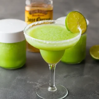 Frozen margarita served in a coupe glass with a salted rim and lime slice on a charcoal stone surface.