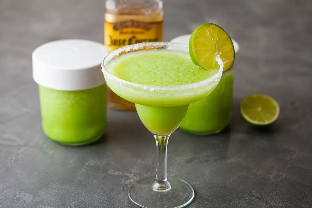 Frozen margarita served in a coupe glass with a salted rim and lime slice on a charcoal stone surface.