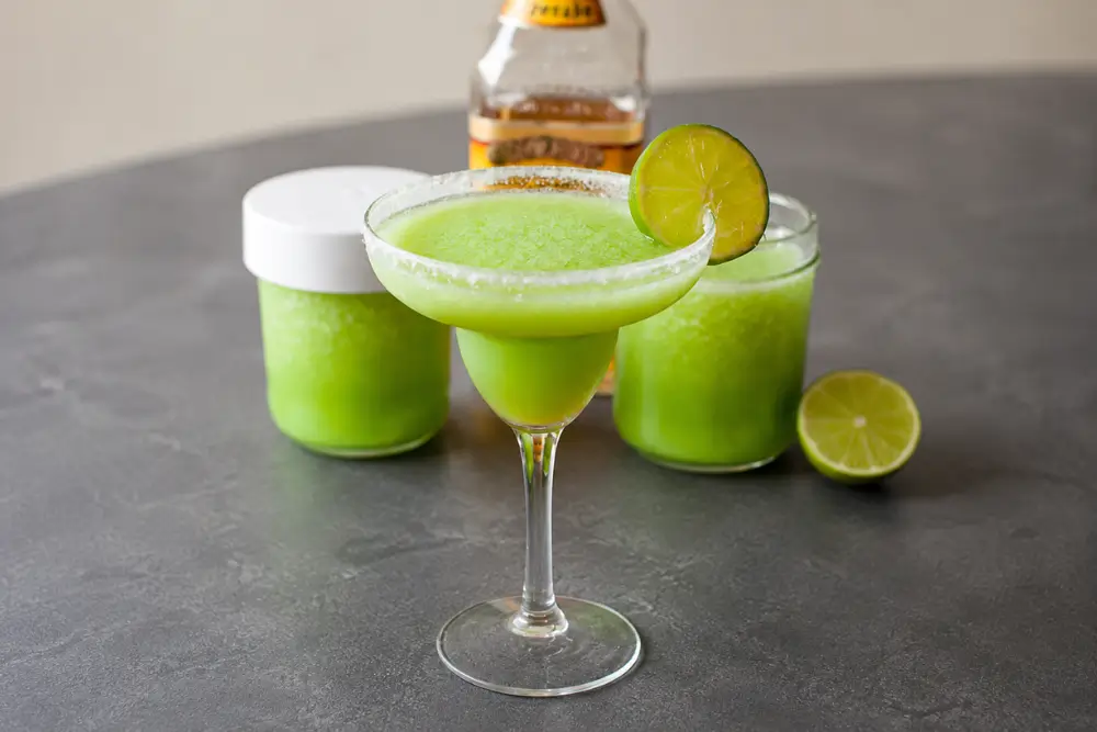 Glass of Make-Ahead Margaritas with a salted rim and lime garnish, served with extra frozen mixture behind it.