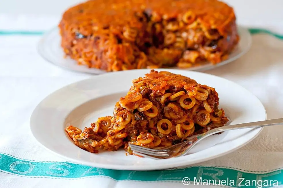 Slice of baked anelletti on a white plate with the pasta bake in the background.