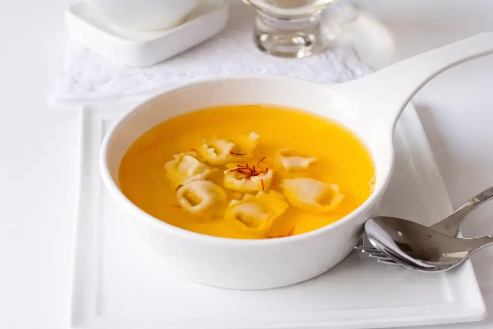 Chicken consommé with saffron served with tortellini in a white bowl.