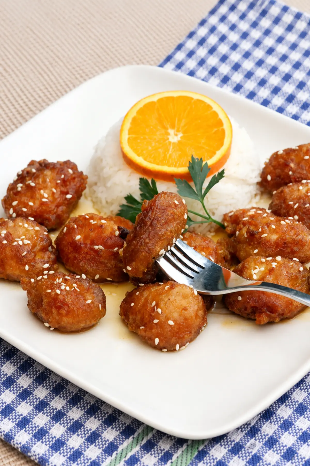 Piece of orange-glazed chicken held on a fork beside steamed rice and sesame-coated chicken pieces.
