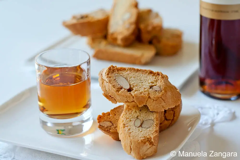 Cantucci slices on a plate beside a glass of sweet wine and a bottle in the background.