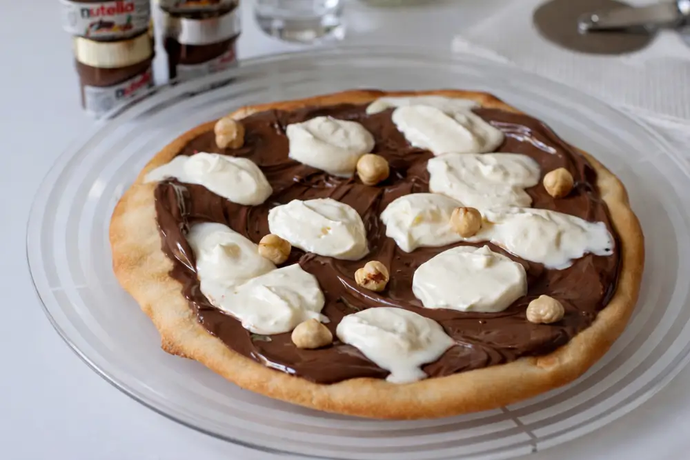Slice of Nutella Pizza topped with whipped cream and toasted hazelnuts on a white plate.