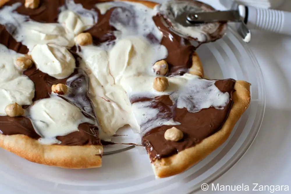 Slice of Nutella Pizza topped with whipped cream and toasted hazelnuts being lifted from a plate.