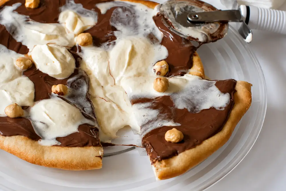 Slice of Nutella Pizza topped with whipped cream and toasted hazelnuts on a white plate.