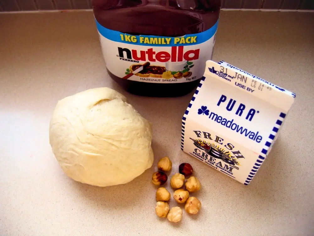 Pizza dough, jar of Nutella, fresh cream and whole hazelnuts arranged on a kitchen counter.