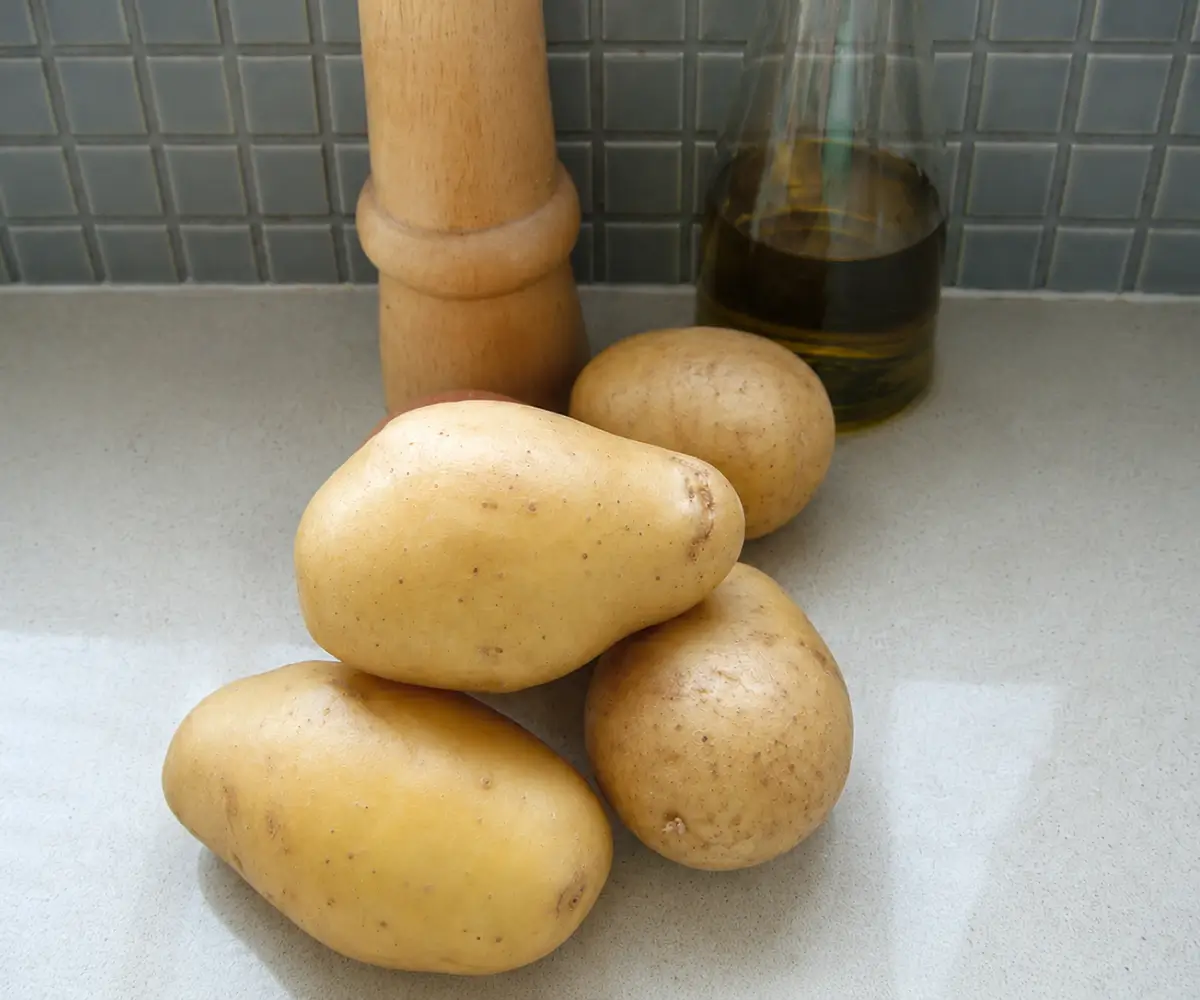 Raw potatoes on a kitchen countertop beside olive oil and a pepper mill.