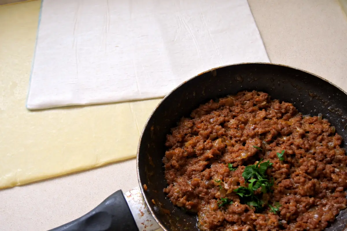 Cooked beef filling in a pan with pastry sheets laid out on the counter.