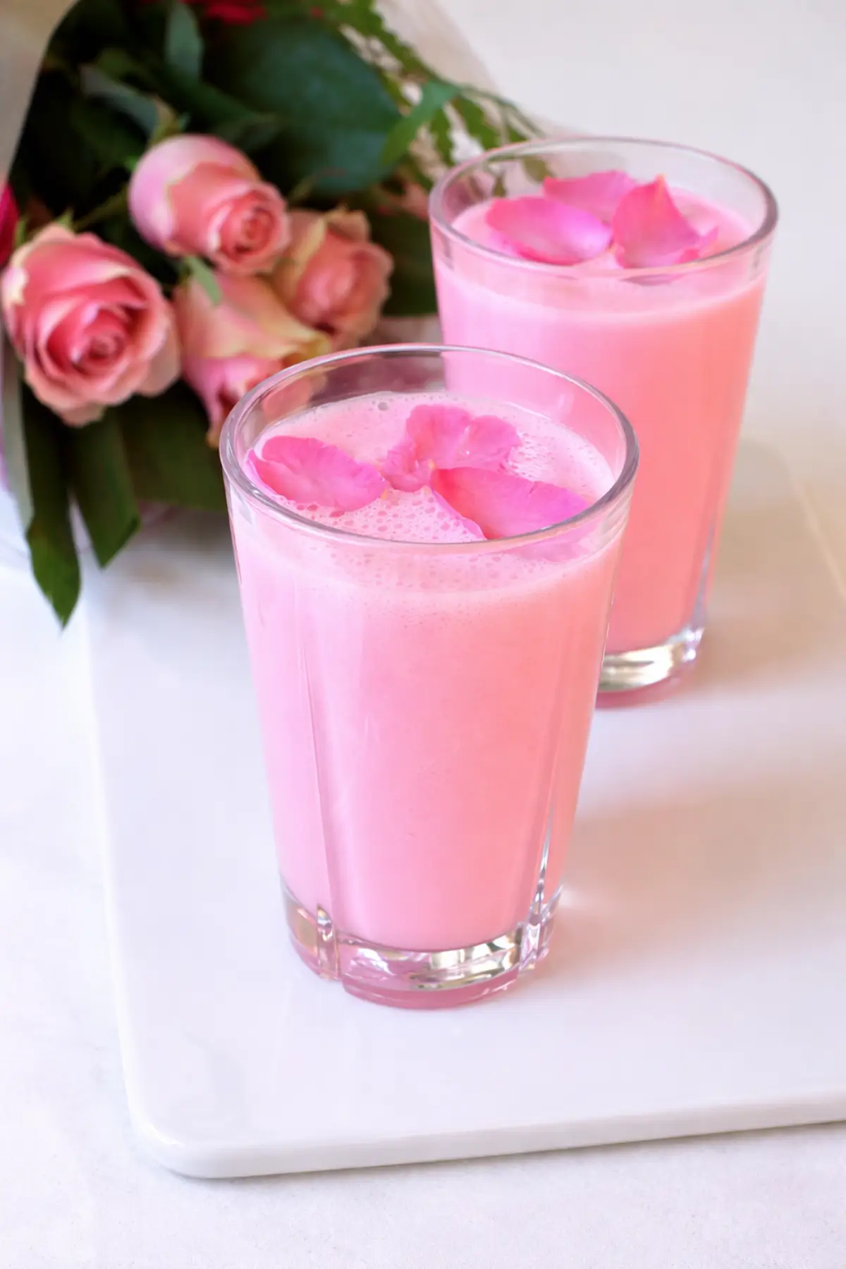 Rose lassi served in short glasses with a bouquet of pink roses beside.