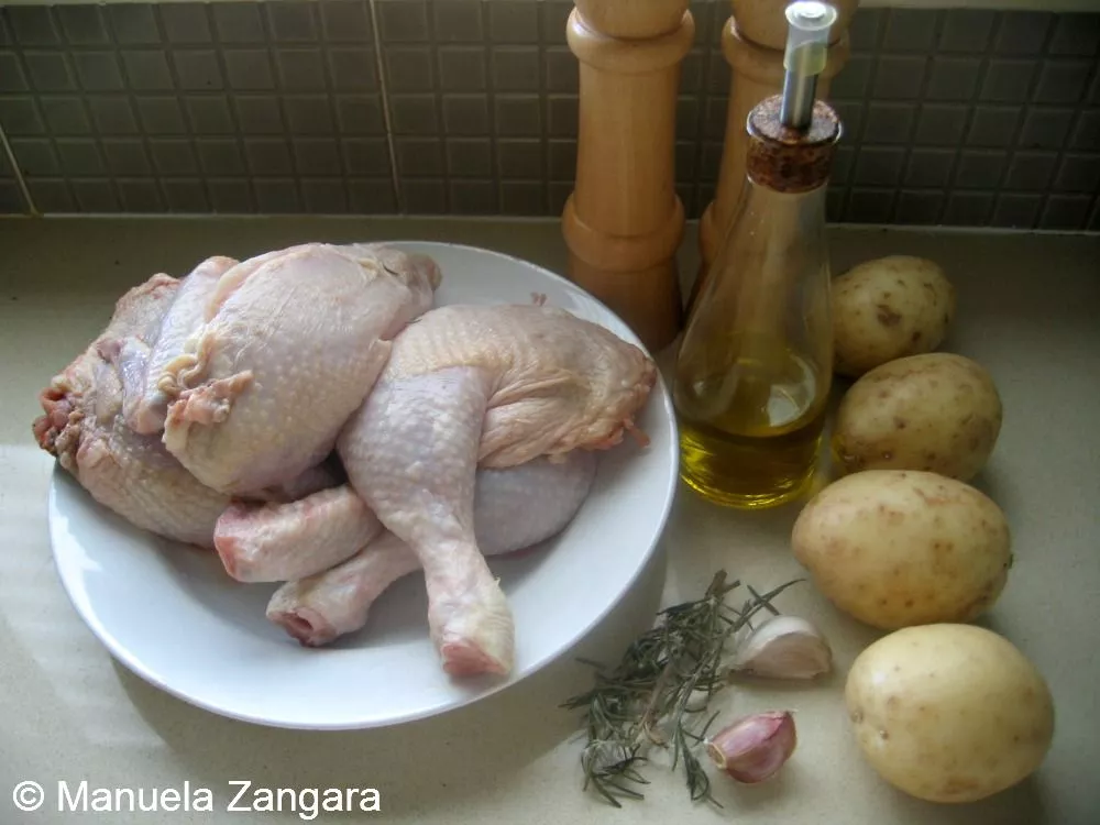 Whole chicken, potatoes, garlic, rosemary, and olive oil arranged on a kitchen counter.