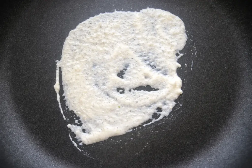 Dosa cooking on a pan with bubbles forming and edges beginning to set.