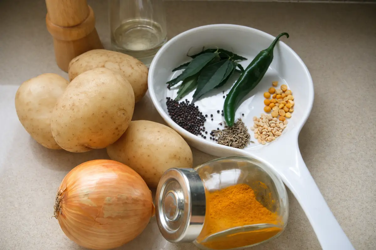 Raw potatoes, onion, green chilli, curry leaves, and whole spices arranged before cooking.