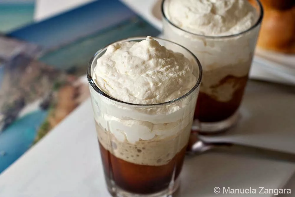 Granita al caffe' con panna Two glasses of coffee granita topped with whipped cream on a white plate beside a spoon.