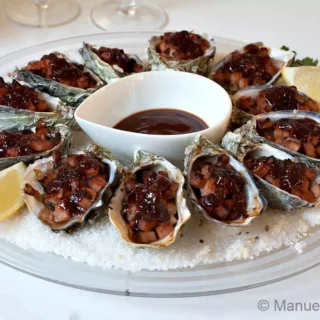 Cooked Oysters Kilpatrick served on a round plate with Kilpatrick sauce and lemon wedges.