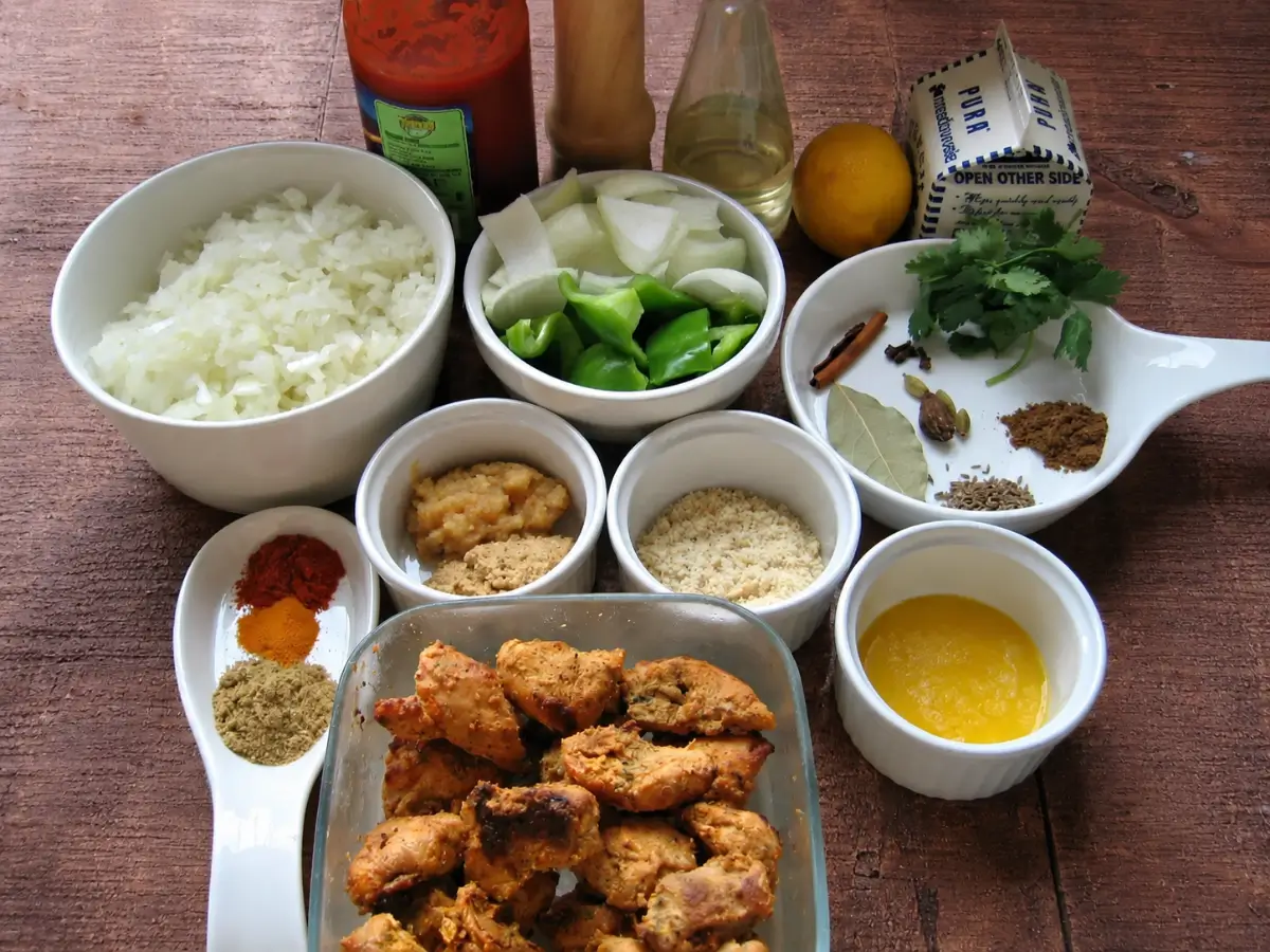 IMG_3039 Ingredients for Chicken Tikka Masala arranged in bowls with spices, chicken tikka, onions, capsicum, butter, lemon, and tomato purée on a rustic wooden table.