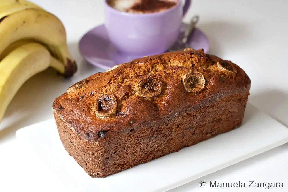 Banana walnut bread loaf topped with banana slices next to fresh bananas and a cappuccino.