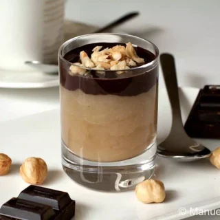 Single glass of pear mousse topped with melted dark chocolate and chopped hazelnuts on a white plate with chocolate squares.