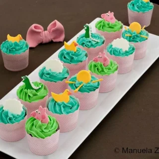A tray of Peppa Pig cupcakes decorated with green and blue icing and fondant figures of Peppa, ducks, and clouds.