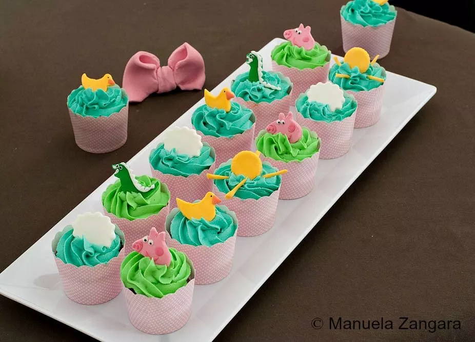 Peppa Pig Chocolate & Vanilla Cupcakes A tray of Peppa Pig cupcakes decorated with green and blue icing and fondant figures of Peppa, ducks, and clouds.