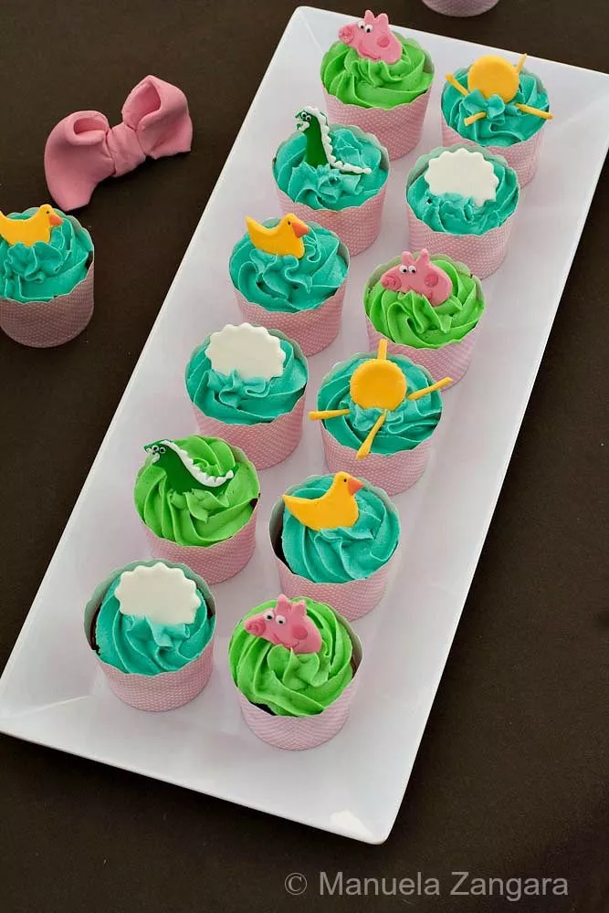Peppa Pig Chocolate & Vanilla Cupcakes Overhead shot of Peppa Pig cupcakes with green and blue icing with fondant toppers of Peppa, dinosaurs, and clouds.