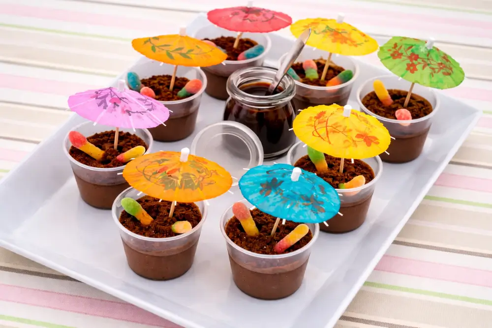 Peppa Pig Muddy Puddle Cups decorated with biscuit crumbs, gummy worms, and colourful paper umbrellas.