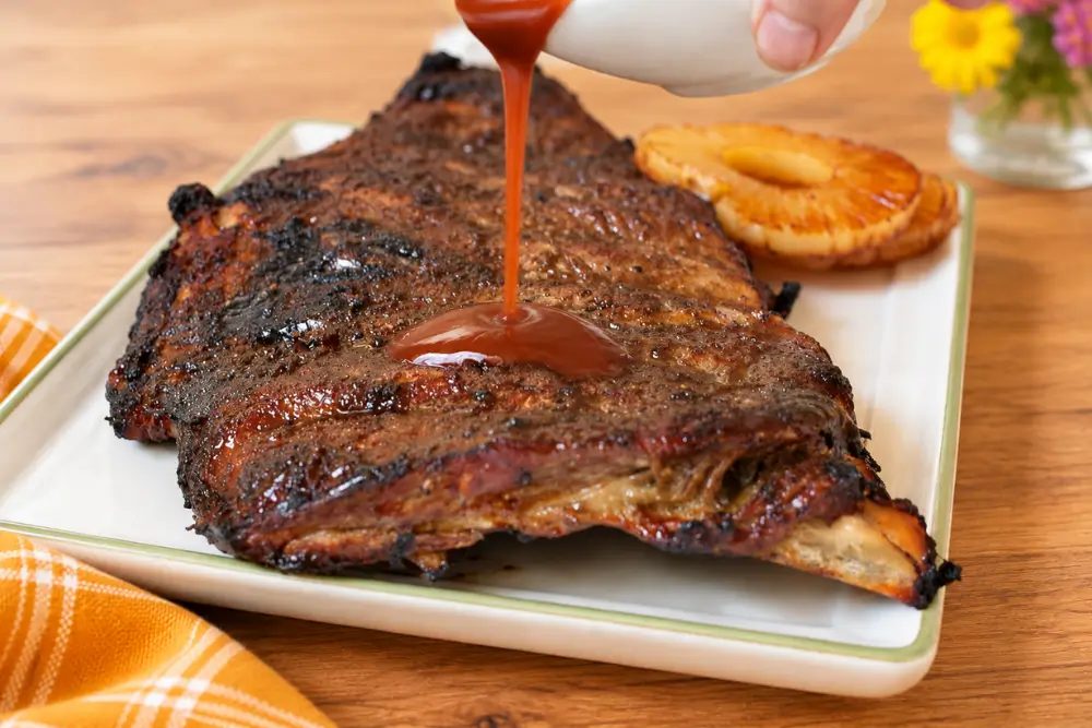 Homemade barbecue sauce being poured over a rack of glazed pork ribs.