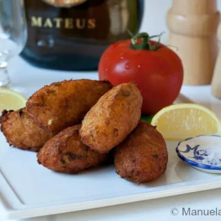 Golden Pasteis de Bacalhau stacked on a plate with lemon wedges and tomato in the background.