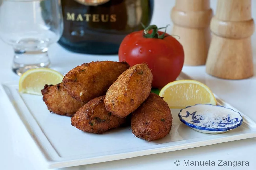 Golden Pasteis de Bacalhau stacked on a plate with lemon wedges and tomato in the background.