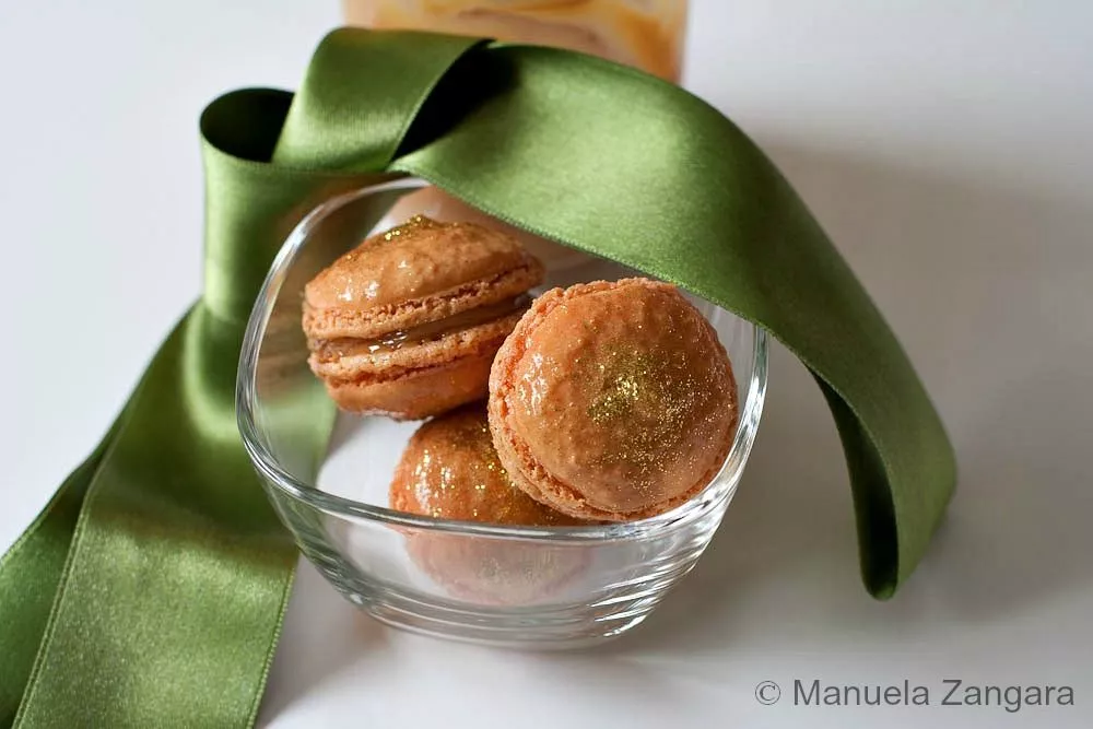 Dulce de Leche macarons in a glass bowl with a soft green ribbon and a jar of caramel in the background.