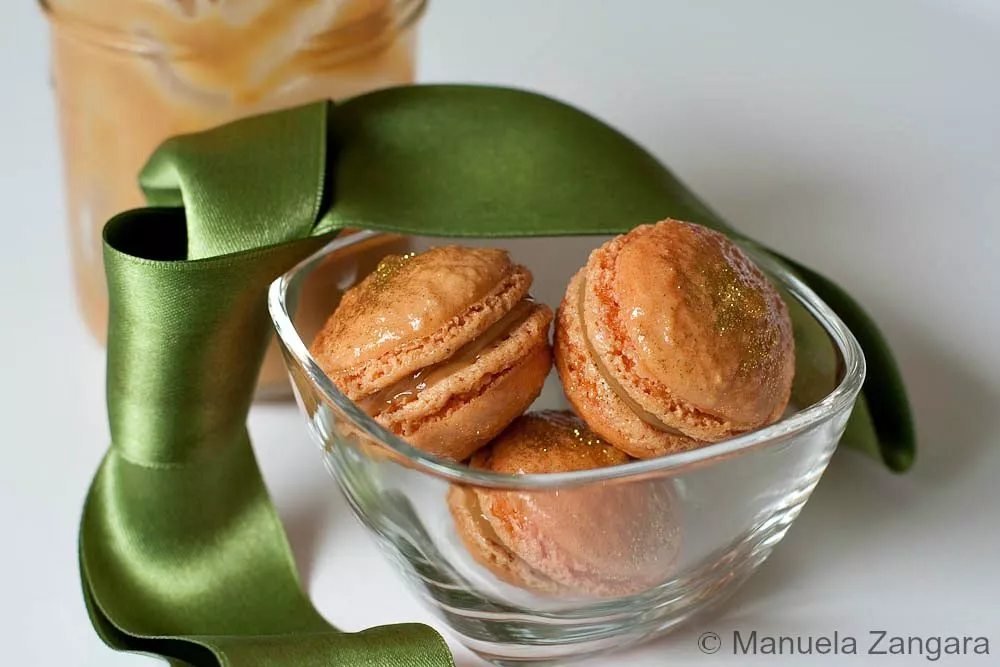 Close-up of Dulce de Leche macarons with gold dust in a glass bowl tied with a green ribbon.