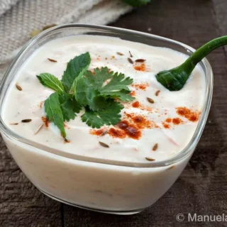 Raita in a glass bowl topped with cumin seeds, coriander, mint, and red chilli powder.