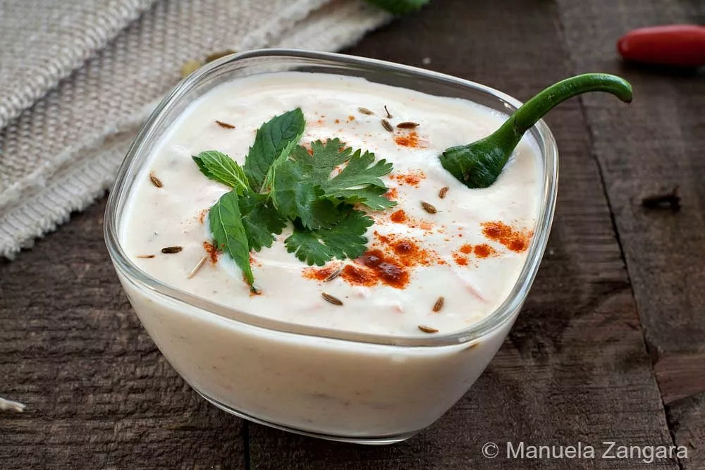 Raita in a glass bowl topped with cumin seeds, coriander, mint, and red chilli powder.