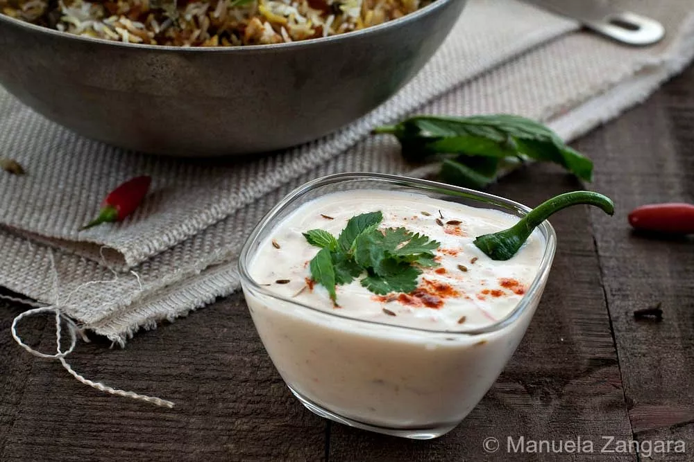A bowl of mixed vegetable raita topped with mint, coriander, and chilli beside a plate of biryani.