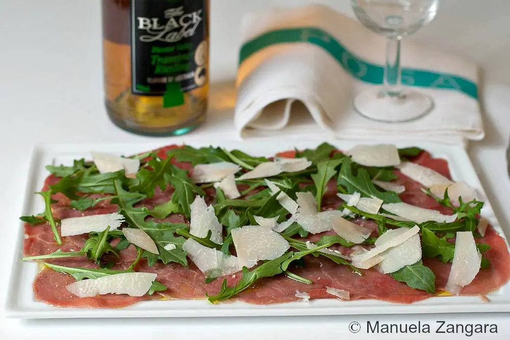 Thin slices of raw beef topped with baby rocket and Grana Padano shavings on a serving plate.