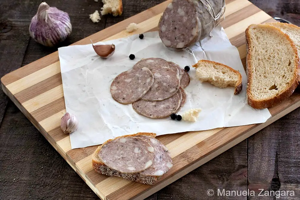 Sliced Salame Cotto arranged on a board with bread, garlic, and peppercorns.