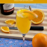 Tall flute filled with Mimosa cocktail and an orange slice on the rim, with oranges and a bottle in the background.