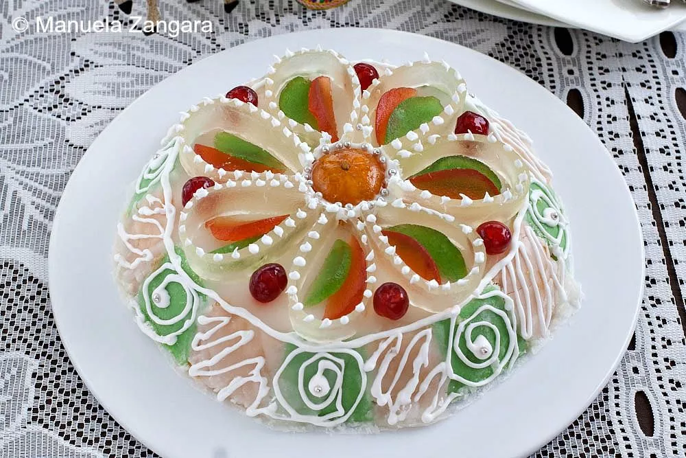 Whole Cassata Siciliana decorated with colorful candied fruits and white icing, placed on a lace tablecloth.