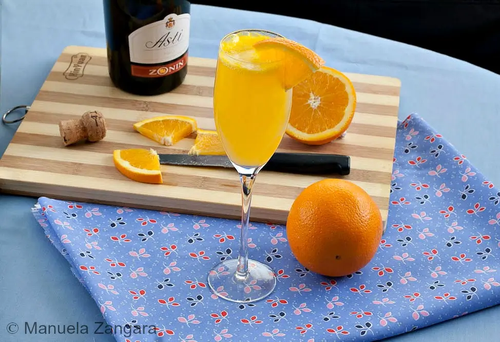 Tall flute filled with Mimosa cocktail and an orange slice on the rim, with oranges and a bottle in the background.