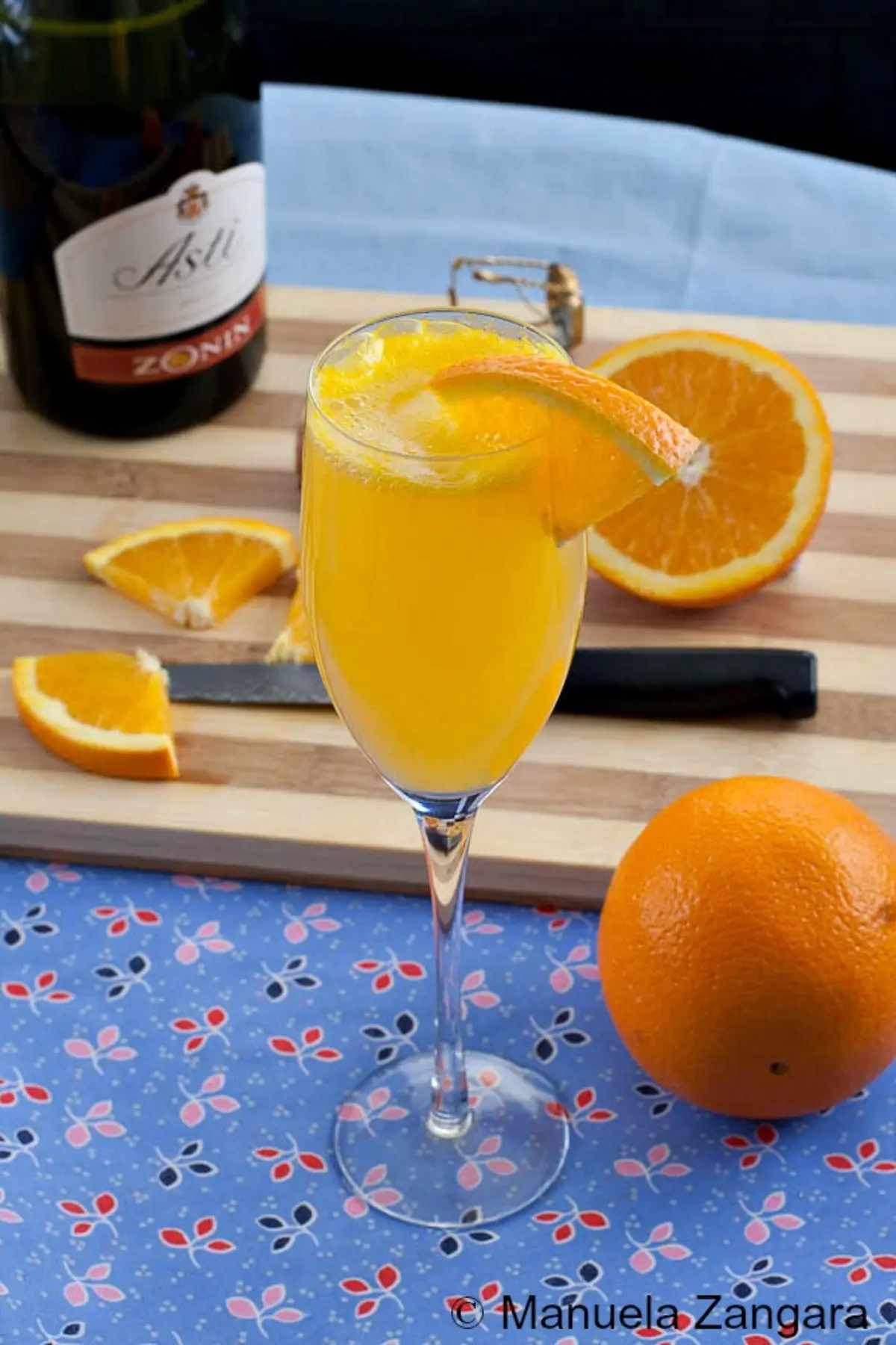 Flute of Mimosa surrounded by fresh oranges, a knife, and a sparkling wine bottle.
